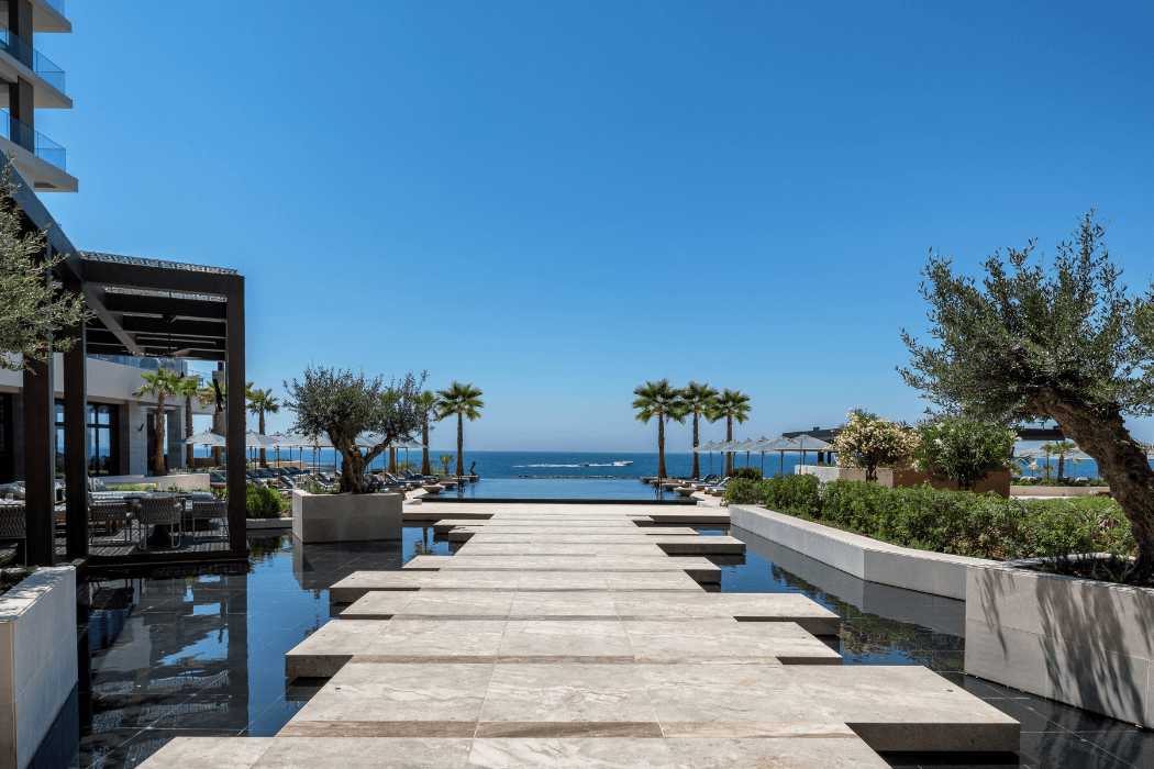 A stone walkway flanked by still reflecting pools leads toward an infinity pool and the open sea. Olive and palm trees, lounge chairs, and shade umbrellas line the terrace under a clear blue sky.