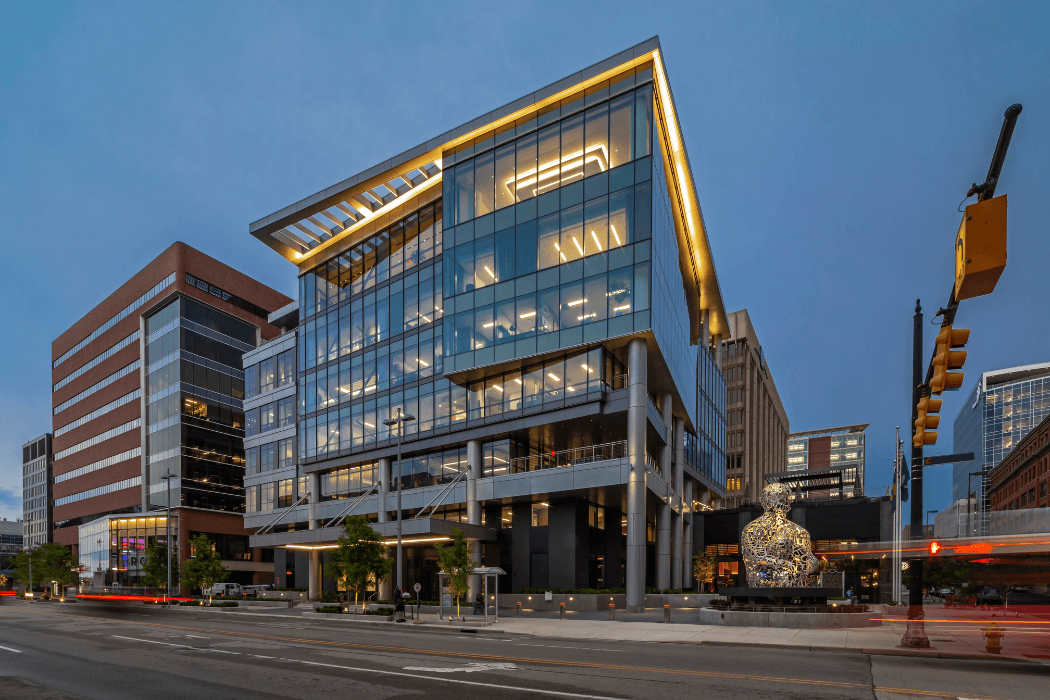 A contemporary multi-story glass office building glows at dusk, its lit interiors and cantilevered upper floor visible. A lattice-work human figure sculpture stands at street level beside brick buildings and traffic light trails.