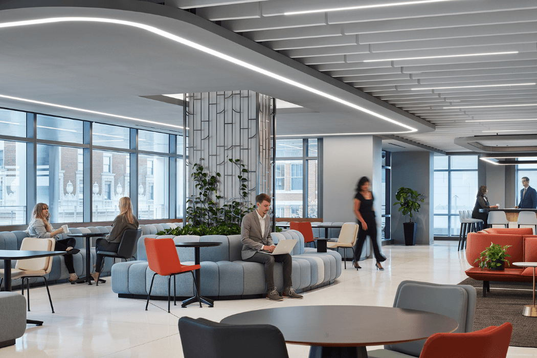 A modern office lounge with a sculptural curved ceiling of recessed linear lights. People work and chat on curved blue banquette seating and bright orange chairs beside floor-to-ceiling windows, with a planted room divider and city buildings visible outside.
