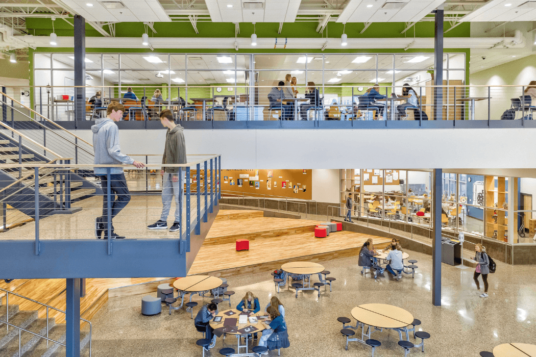 A two-story school commons where students study at round tables on the main floor while others work in classrooms behind glass railings on the mezzanine above. Bright green accent walls and a wood-floored tiered platform anchor the space.