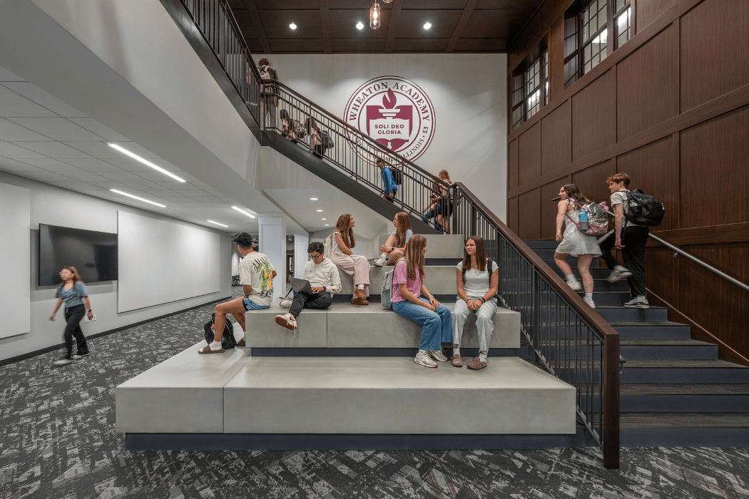 Students gather on tiered concrete bleacher seating at the base of a staircase in a school commons. A maroon Wheaton Academy crest reading "Soli Deo Gloria" is painted on the wall above, next to dark wood paneling.