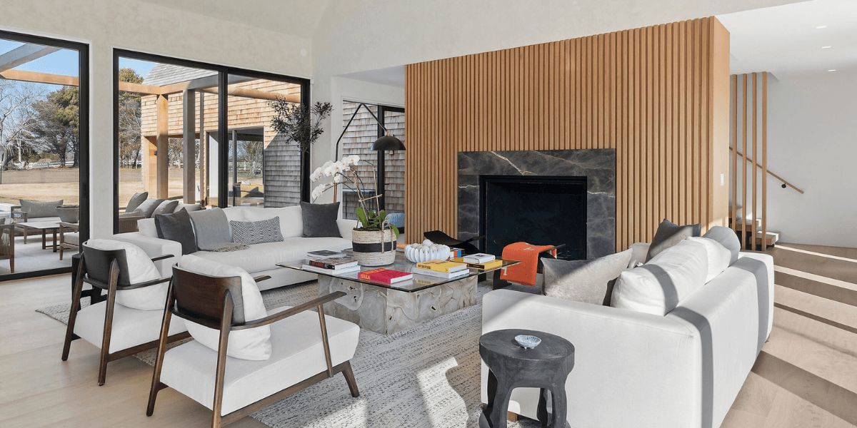A bright, modern living room with two white sofas and a pair of wood-framed armchairs arranged around a stone-topped coffee table on a textured gray rug.